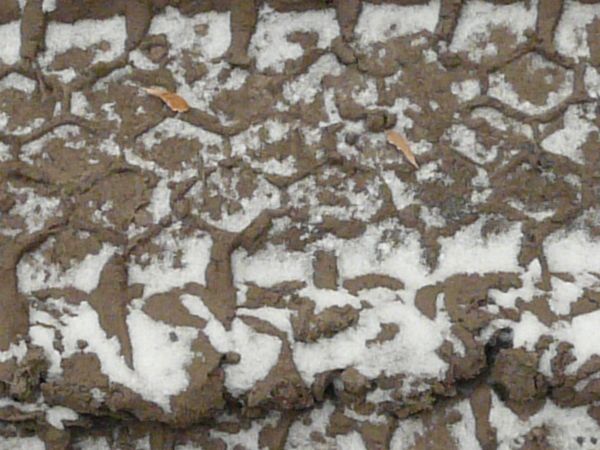 Dirt road texture in dark brown tone with tire marks and thin layer of snow across surface.